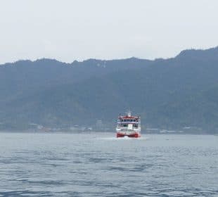 Eine rot-weiße Fähre gleitet über ruhiges Wasser auf die Kamera zu, eingerahmt von bewaldeten Bergen und einem wolkenverhangenen Himmel im Hintergrund.