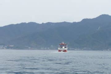 Eine rot-weiße Fähre gleitet über ruhiges Wasser auf die Kamera zu, eingerahmt von bewaldeten Bergen und einem wolkenverhangenen Himmel im Hintergrund.