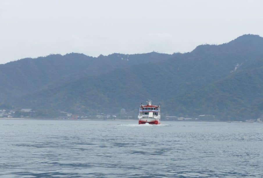 Eine rot-weiße Fähre gleitet über ruhiges Wasser auf die Kamera zu, eingerahmt von bewaldeten Bergen und einem wolkenverhangenen Himmel im Hintergrund.