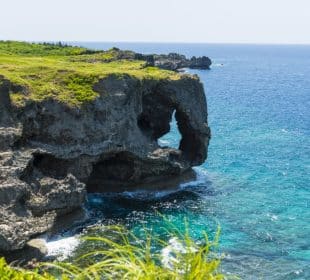 Eine felsige Klippe in Okinawa mit einem natürlichen Bogen, der wie ein Elefantenrüssel geformt ist, erstreckt sich über das klare blaue Wasser des Ozeans, mit grünem Gras auf der Oberseite und Wellen, die an einem sonnigen Tag gegen die Felsen darunter schlagen.