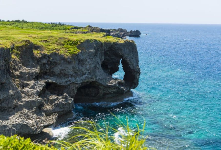 Eine felsige Klippe in Okinawa mit einem natürlichen Bogen, der wie ein Elefantenrüssel geformt ist, erstreckt sich über das klare blaue Wasser des Ozeans, mit grünem Gras auf der Oberseite und Wellen, die an einem sonnigen Tag gegen die Felsen darunter schlagen.