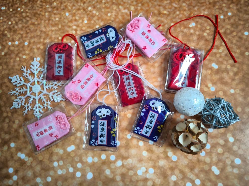 Verschiedene japanische Omamori-Amulette in Rosa und Marineblau sind auf einer glitzernden Oberfläche arrangiert, die den Charme eines Adventskalenders versprüht. In der Nähe befinden sich ein weißes Schneeflocken-Ornament, ein Garnknäuel, eine weiße Kugel und getrocknete Blumenschoten.