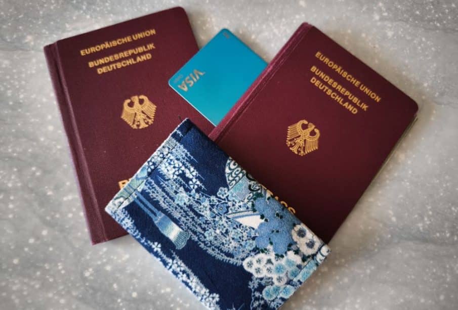 Zwei deutsche Reisepässe, eine blaue Visakarte und ein blaues, blumengemustertes Passetui - möglicherweise verloren - sind auf einer hellgrauen, gesprenkelten Oberfläche angeordnet.