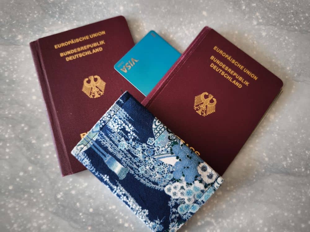 Zwei deutsche Reisepässe, eine blaue Visakarte und ein blaues, blumengemustertes Passetui - möglicherweise verloren - sind auf einer hellgrauen, gesprenkelten Oberfläche angeordnet.
