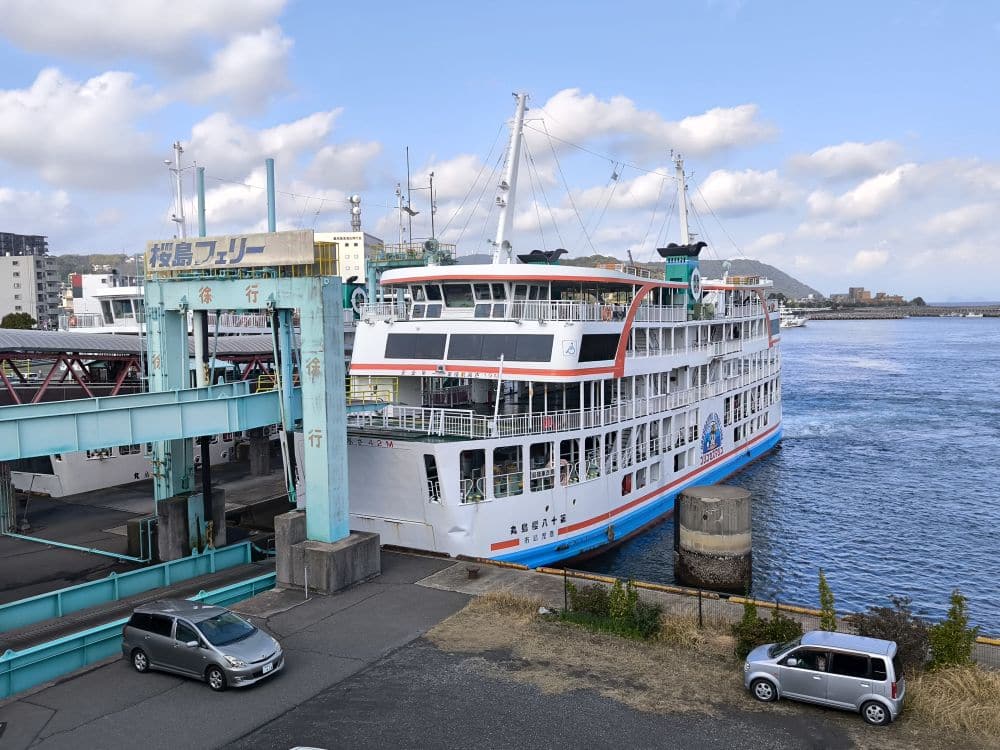 Eine große weiße Fähre liegt bei bewölktem Himmel an einem Hafenterminal an, mit einer Rampe, die die Fähre mit dem Terminal verbindet. Zwei geparkte Autos sind auf der Straße in der Nähe des Ufers zu sehen.