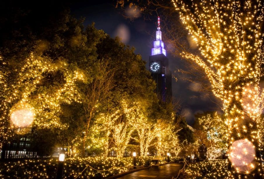 Eine nächtliche Szene mit Bäumen, die mit goldenen Illuminationen geschmückt sind und einen Weg säumen, während im Hintergrund ein hoher Uhrenturm in Lila und Weiß leuchtet. Lichtreflexe und Regentropfen tragen zu dem verträumten Effekt bei.