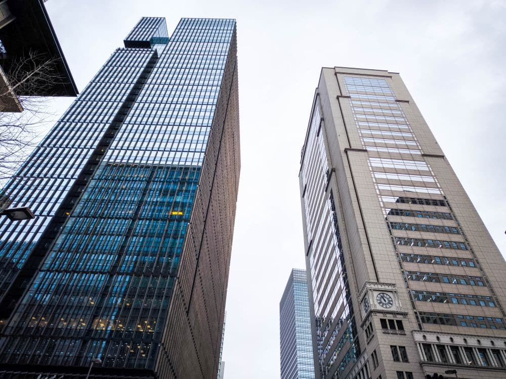 Zwei hohe Bürogebäude mit Glas- und Metallfassaden stehen nebeneinander in Marunouchi, vom Straßenniveau aus gesehen. Das Gebäude auf der rechten Seite hat eine Uhr über seinem Eingang. Der Himmel ist bedeckt.