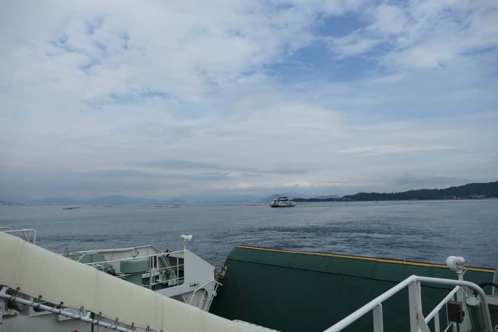 Blick von einem Boot aus über ruhiges Wasser mit einer fernen Fähre am Horizont; teilweise bewölkter Himmel und Land in der Ferne.