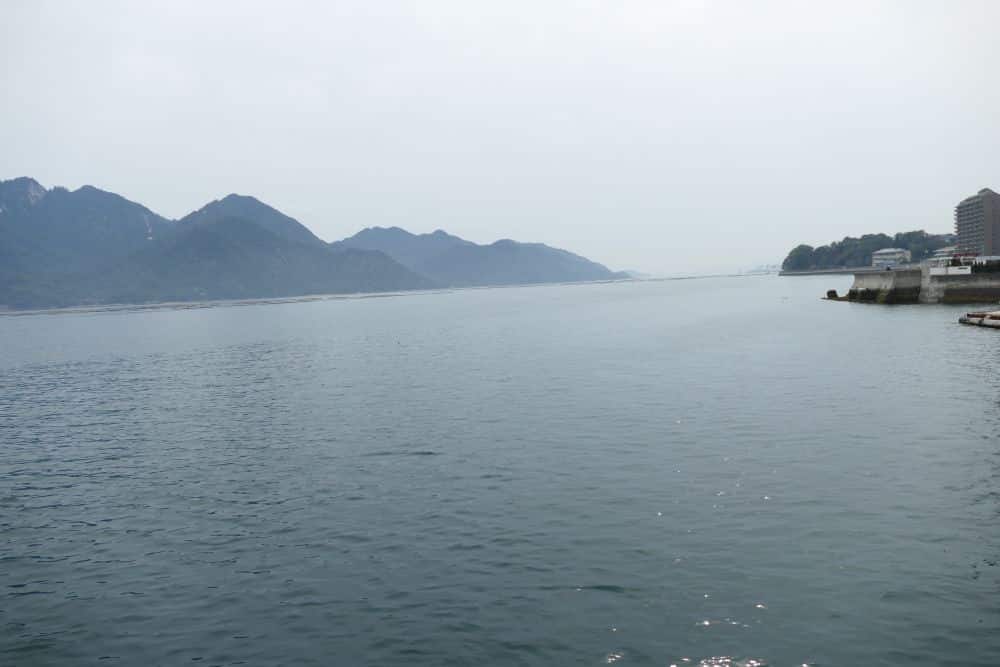 Ein ruhiges, weites Gewässer mit fernen Bergen unter einem dunstigen Himmel. Auf der rechten Seite säumen Gebäude und ein kleiner Steg das Ufer, während eine Fähre ruhig über die sanfte Oberfläche gleitet.
