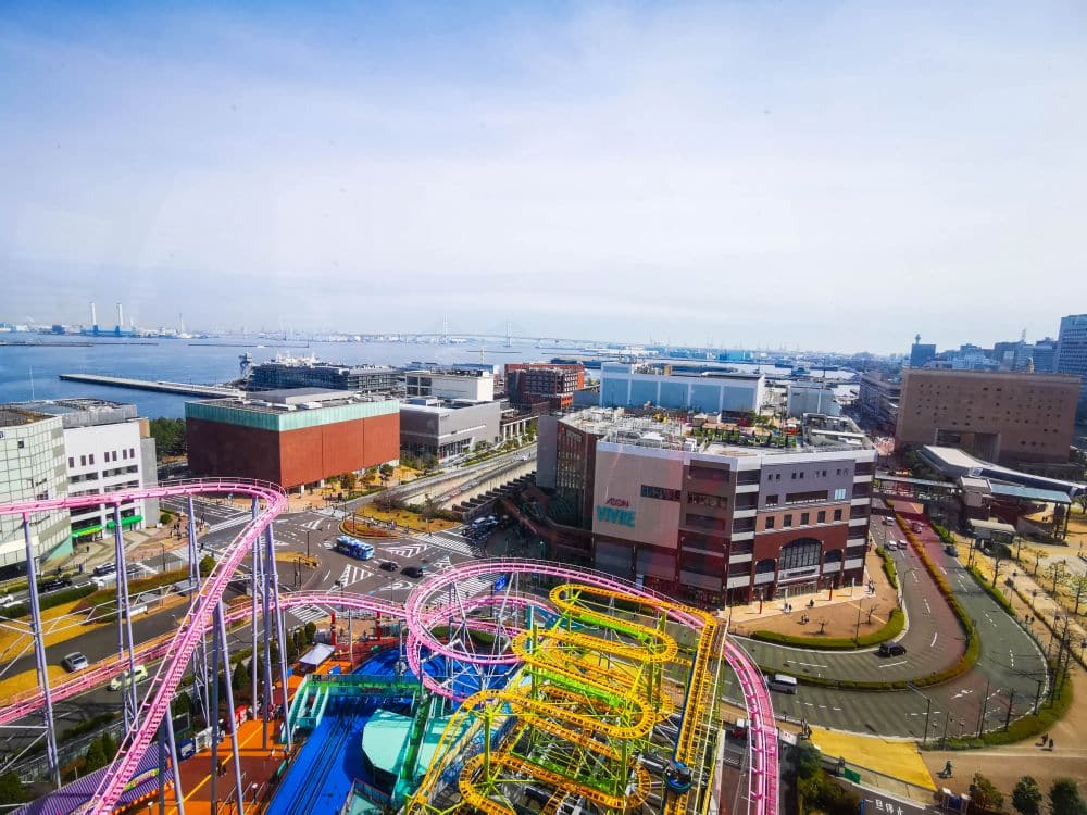 Luftaufnahme einer Küstenstadt mit den bunten Fahrgeschäften des Cosmo World-Vergnügungsparks im Vordergrund, modernen Gebäuden, Straßen und einer Brücke, die sich unter einem klaren Himmel über das Wasser erstreckt.