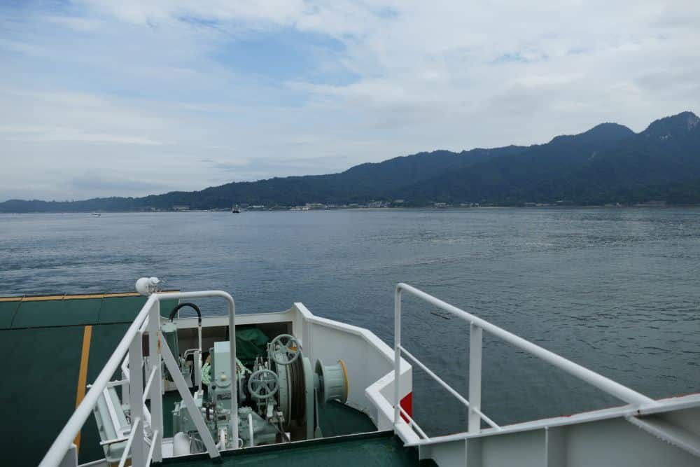 Blick vom Deck einer Fähre über ruhiges Wasser auf eine entfernte Küste mit Bergen und einem bewölkten Himmel im Hintergrund. Im vorderen Teil des Schiffes sind einige Maschinen und die Reling zu sehen.