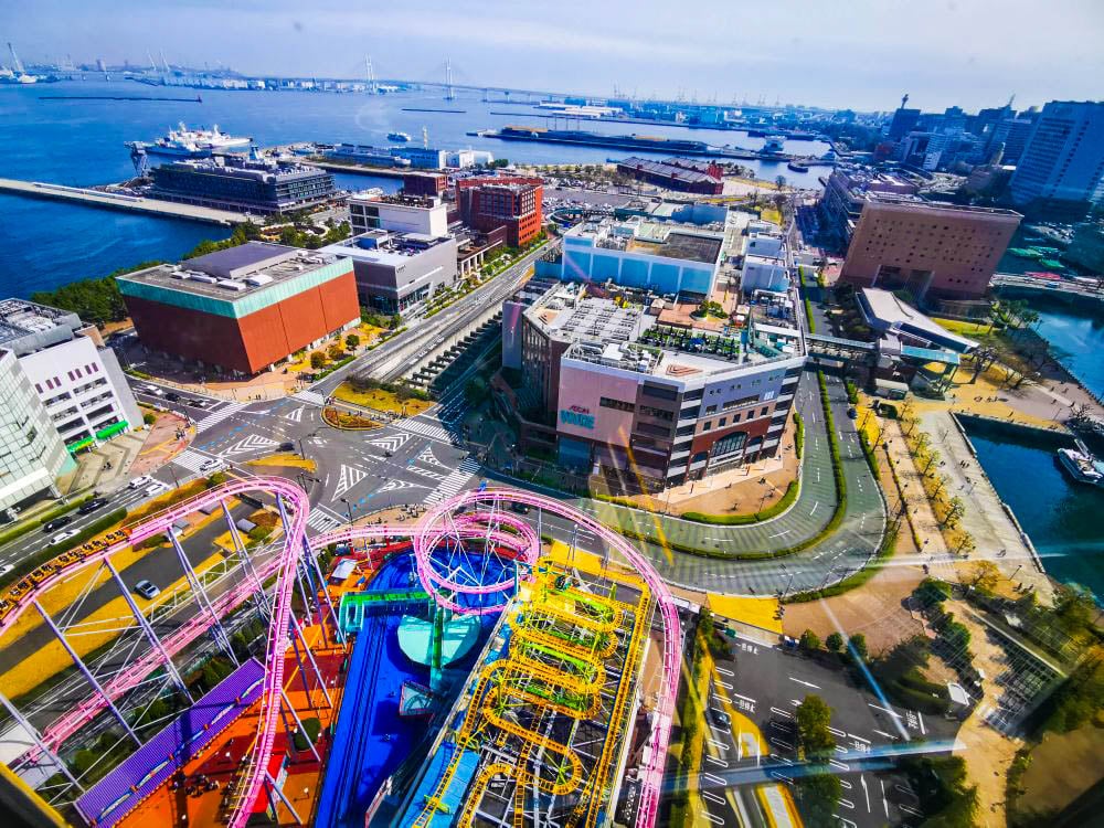 Ein pulsierendes Stadtbild mit dem farbenfrohen Vergnügungspark Cosmo World und seinen aufregenden Achterbahnen im Vordergrund, modernen Gebäuden, Straßen und einem Hafen mit Schiffen unter einem klaren blauen Himmel.