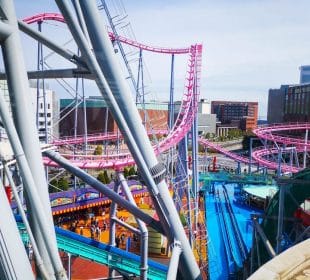 Der lebhafte Vergnügungspark Cosmo World bietet gewundene rosa Achterbahnschienen über bunten Attraktionen und Gebäuden, unter denen die Besucher an einem sonnigen Tag spazieren gehen können.