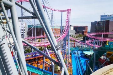 Der lebhafte Vergnügungspark Cosmo World bietet gewundene rosa Achterbahnschienen über bunten Attraktionen und Gebäuden, unter denen die Besucher an einem sonnigen Tag spazieren gehen können.