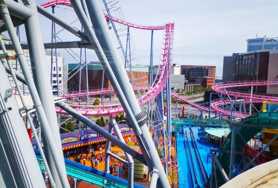 Der lebhafte Vergnügungspark Cosmo World bietet gewundene rosa Achterbahnschienen über bunten Attraktionen und Gebäuden, unter denen die Besucher an einem sonnigen Tag spazieren gehen können.