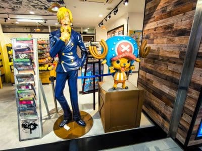 Lebensgroße Statuen von Sanji und Chopper, den beliebten Anime-Figuren aus One Piece, stehen in einem hell erleuchteten Laden in Tokio, während im Hintergrund Regale mit Manga und Merchandise-Artikeln aufgebaut sind.