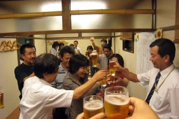 Eine Gruppe von Menschen in einem Innenraum erhebt Gläser mit Bier und lächelt, als würde sie gemeinsam bei einem lebhaften Bonenkai feiern. Einige reichen sich über einen Tisch hinweg die Hände, und die Atmosphäre ist fröhlich und festlich.