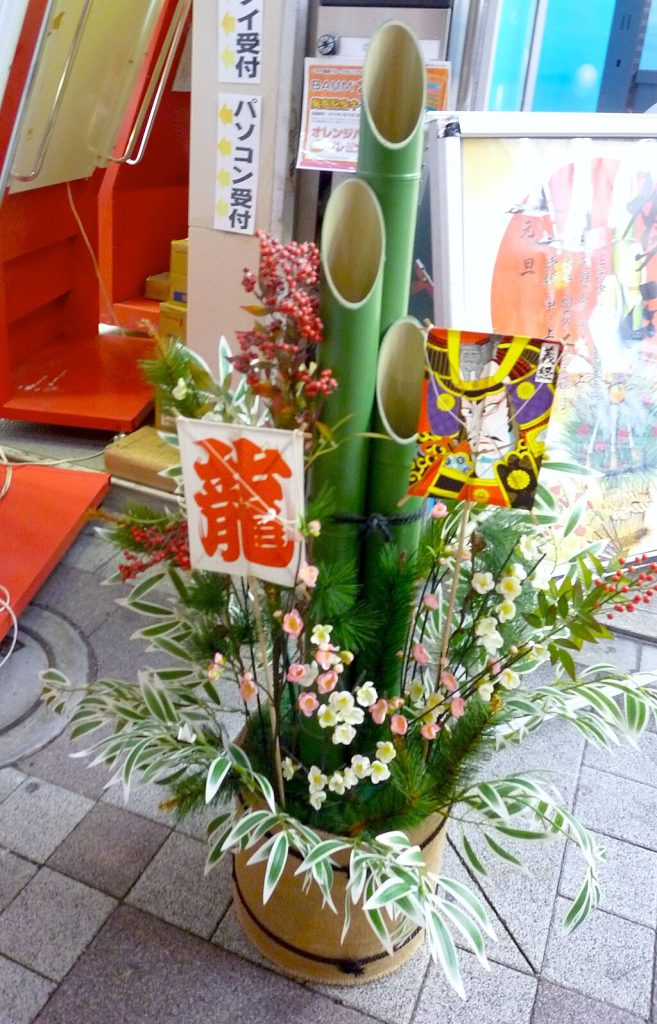 Ein dekoratives Kadomatsu-Arrangement mit Bambus, Kiefer, Blumen und festlichen Karten steht auf dem Bürgersteig vor einem Geschäft.