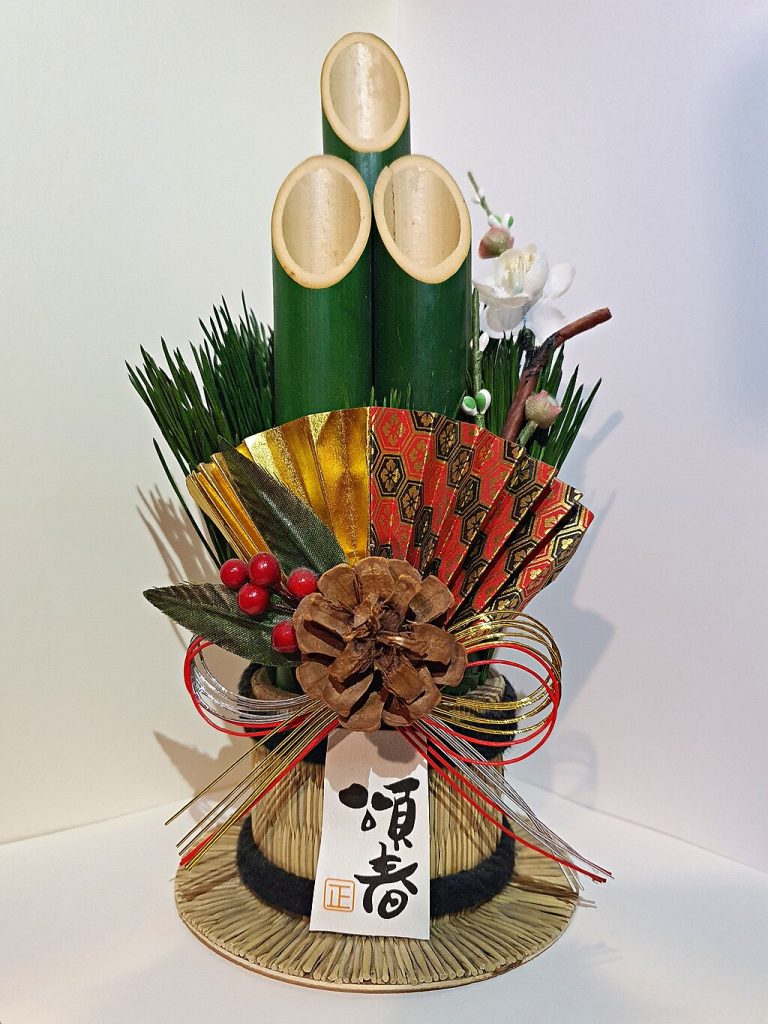 Ein dekoratives Kadomatsu-Arrangement für das japanische Neujahrsfest mit drei Bambushalmen, Kiefernzweigen, roten Beeren, einem Kiefernzapfen, einem verzierten Fächer und einem weißen Anhänger mit japanischer Schrift, alles auf einem Strohsockel.