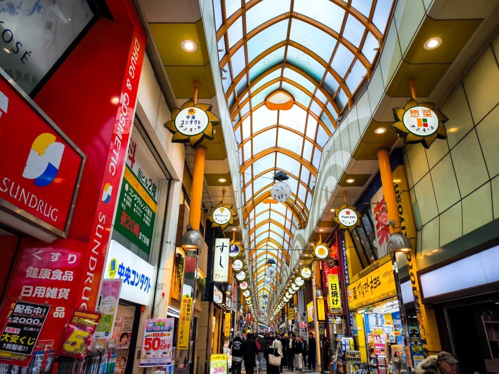 Eine belebte überdachte Einkaufsstraße in der Nähe des Nakano Broadway in Japan mit bunten Schildern, hängenden Laternen und Menschen, die unter einem hohen Bogendach spazieren gehen. Die Geschäfte sind hell erleuchtet und dicht aneinander gedrängt, was eine lebendige, pulsierende Atmosphäre schafft.