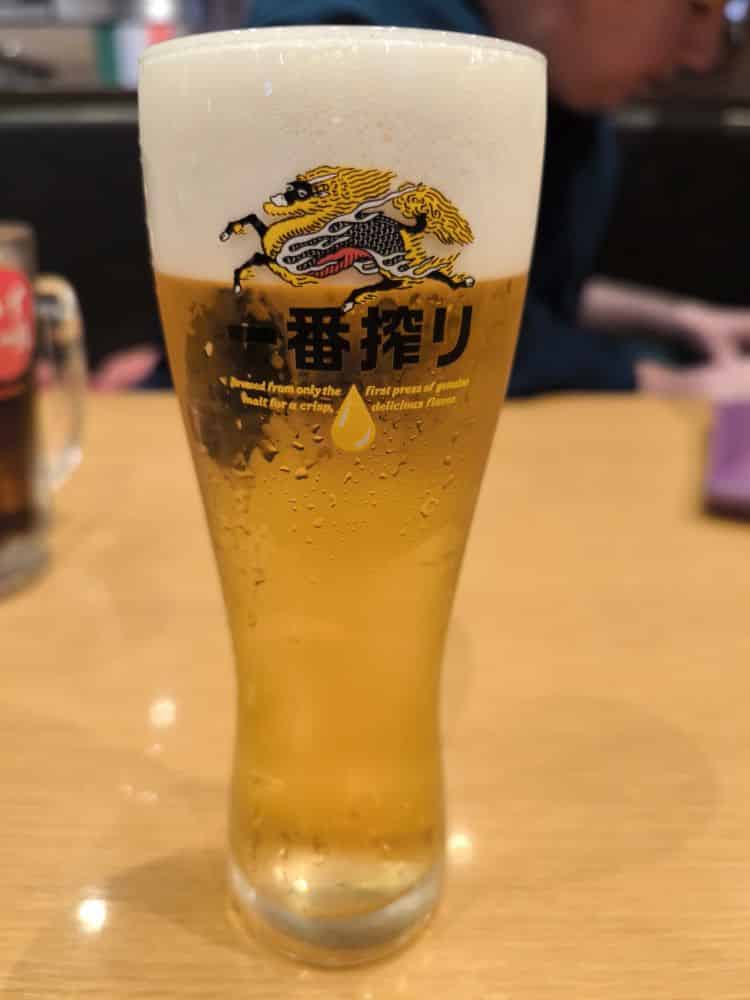 Ein hohes Glas helles Bier mit Schaumkrone steht auf einem Holztisch. Auf dem Glas ist das Logo von Kirin Ichiban mit einem Drachen abgebildet, perfekt für die Feier des Bonenkai, während sich im verschwommenen Hintergrund Menschen versammeln.