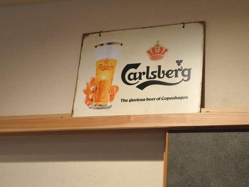 Ein altes Carlsberg-Schild mit einer Hand, die ein Glas Bier hält, und dem Text Carlsberg und Das glorreiche Bier von Kopenhagen, perfekt für Bonenkai-Treffen, an einer beigen Wand über einem Holzregal.