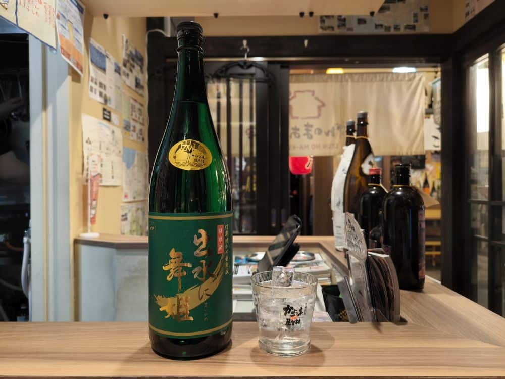 Eine große grüne Flasche mit japanischem Schriftzug steht auf einem Holztresen neben einem klaren Glas mit Flüssigkeit - und fängt die Essenz einer Bonenkai-Feier im warm beleuchteten Inneren eines japanischen Restaurants oder einer Bar ein.