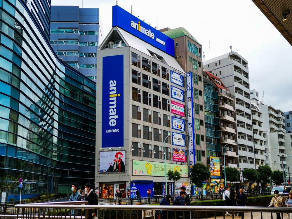 Straßenansicht von Ikebukuro, einem belebten Stadtviertel in Japan, das urbanen Charme mit modernen Gebäuden, auffälligen belebten Schildern, lebhafter Anime-Kultur-Werbung und maskierten Fußgängern auf dem belebten Bürgersteig verbindet.