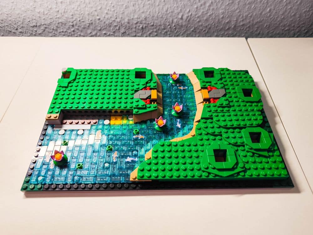 Eine von einem Garten inspirierte LEGO Landschaft mit grünen Hügeln, einem blauen Fluss aus transparenten Steinen, kleinen gelben und rosafarbenen Blumen und einem beigen Weg, der sich auf einer flachen weißen Fläche durch die Landschaft schlängelt.