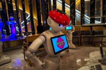 Ein humanoider Roboter, der Pepper ähnelt, trägt drinnen einen großen Erdbeerhut und zeigt auf seinem Brustbildschirm japanischen Text an. Im Hintergrund sind stilvolle Sitzgelegenheiten, beleuchtete Rolltreppen und ein weiterer Roboter in blauer Beleuchtung zu sehen.