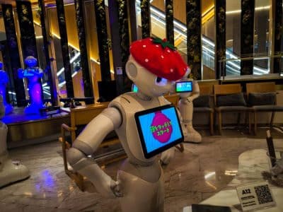 Ein humanoider Roboter, der Pepper ähnelt, trägt drinnen einen großen Erdbeerhut und zeigt auf seinem Brustbildschirm japanischen Text an. Im Hintergrund sind stilvolle Sitzgelegenheiten, beleuchtete Rolltreppen und ein weiterer Roboter in blauer Beleuchtung zu sehen.