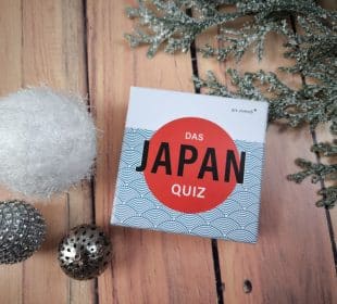 Eine Schachtel mit der Aufschrift "Das Japan-Quiz" steht auf einer hölzernen Unterlage neben einem weißen Fuzzy-Ball, zwei metallischen Zierkugeln und einem mattierten immergrünen Zweig.