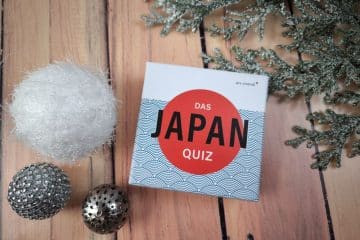 Eine Schachtel mit der Aufschrift "Das Japan-Quiz" steht auf einer hölzernen Unterlage neben einem weißen Fuzzy-Ball, zwei metallischen Zierkugeln und einem mattierten immergrünen Zweig.