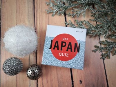 Eine Schachtel mit der Aufschrift "Das Japan-Quiz" steht auf einer hölzernen Unterlage neben einem weißen Fuzzy-Ball, zwei metallischen Zierkugeln und einem mattierten immergrünen Zweig.