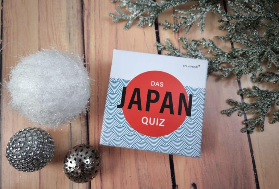 Eine Schachtel mit der Aufschrift "Das Japan-Quiz" steht auf einer hölzernen Unterlage neben einem weißen Fuzzy-Ball, zwei metallischen Zierkugeln und einem mattierten immergrünen Zweig.