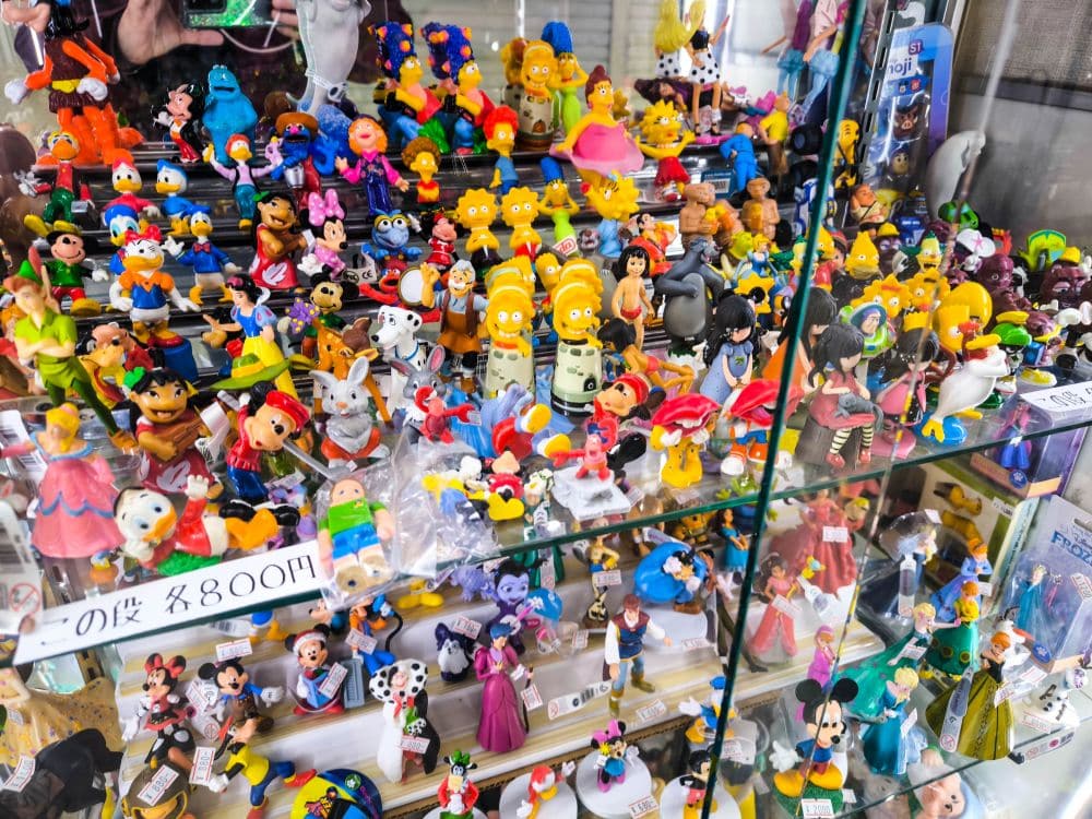 Eine Glasvitrine mit farbenfrohen Comic- und Zeichentrickfiguren, darunter die Simpsons und die Muppets, mit Preisschildern in japanischen Yen, die an die lebhaften Sammlerläden am Nakano Broadway erinnern.