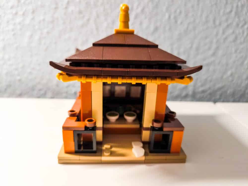 Ein kleines LEGO-Modell eines Gebäudes im asiatischen Stil mit einem braunen, geschwungenen Dach, gelben und orangefarbenen Wänden und zwei kleinen runden Objekten im Inneren, die an einen ruhigen Garten erinnern, auf einer weißen Fläche mit grauem Hintergrund.