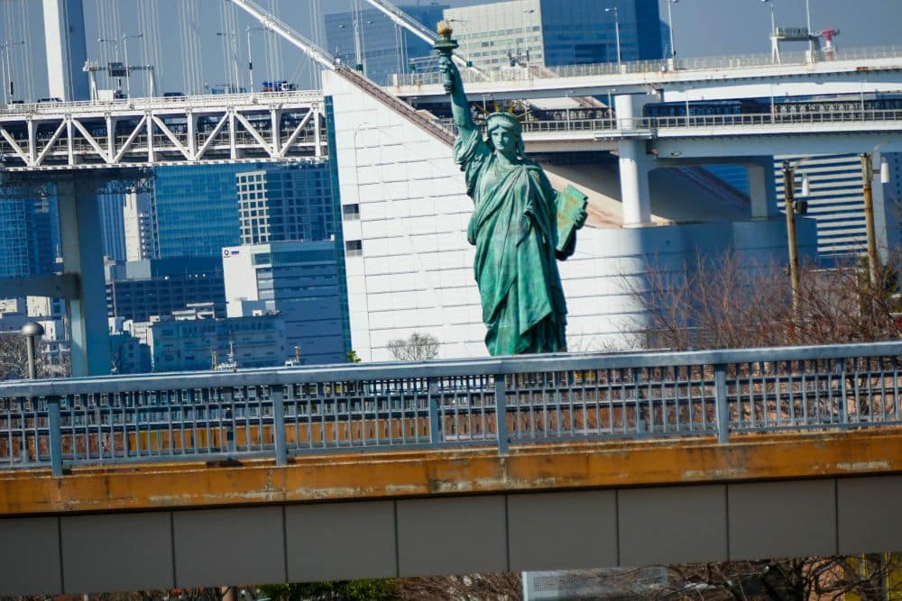 Eine Nachbildung der Freiheitsstatue in Tokio steht an einem sonnigen Tag auf einer Plattform mit städtischen Gebäuden und einem Netz von Brücken im Hintergrund.