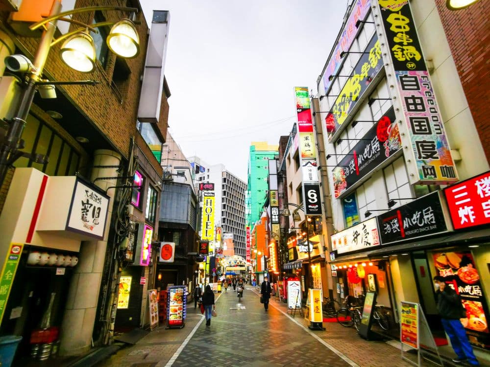 Eine belebte Straßenszene in Ikebukuro, Japan, mit leuchtenden Neonschildern und hohen Gebäuden, fängt den urbanen Charme der Menschen ein, die entlang einer belebten Fußgängerzone mit Geschäften und Restaurants flanieren.