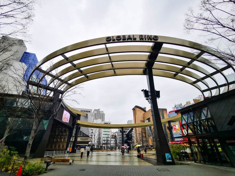 Ein großer, moderner Rundbau mit der Aufschrift GLOBAL RING steht auf einem offenen Platz in Ikebukuro. An einem bewölkten Tag spazieren die Menschen unter ihm hindurch, umgeben von Gebäuden, Bäumen, digitalen Bildschirmen und dem urbanen Charme der Anime-Kultur.