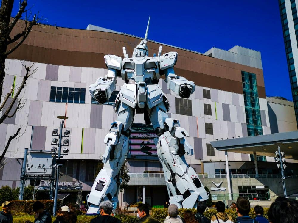 Die majestätische Einhorn-Gundam-Statue ragt vor einem modernen Gebäude in Odaiba, Tokio, in die Höhe, und die Menschen versammeln sich unter einem strahlend blauen Himmel an ihrem Sockel.