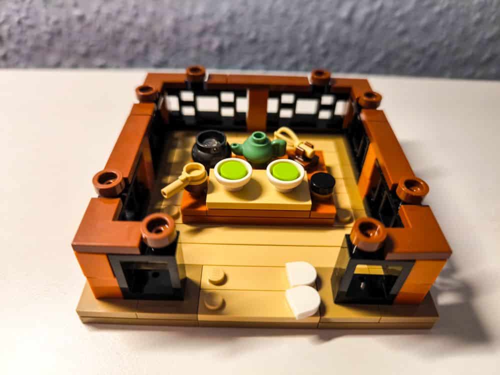 Ein kleines Lego-Modell einer Teestube mit braunen Wänden blickt auf einen charmanten Garten mit zwei weißen Stühlen, einem Tisch mit grünen Tassen, einer Teekanne, einer Schale und verschiedenen Utensilien, die alle auf einer weißen Fläche stehen.