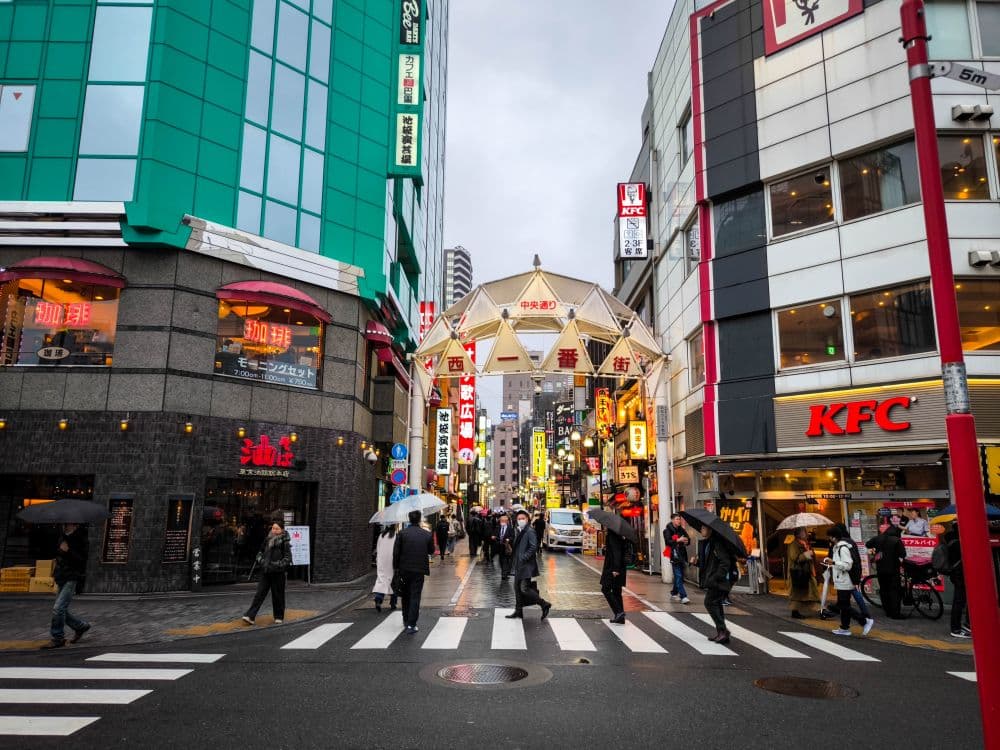 Menschen mit Regenschirmen überqueren eine Straße an einer belebten Ikebukuro-Kreuzung, die von Geschäften und Restaurants, darunter ein KFC, gesäumt ist. Helle Schilder und Lichter fangen den urbanen Charme ein, mit einem überdachten Eingang einer Einkaufspassage im Hintergrund an einem bewölkten Tag.