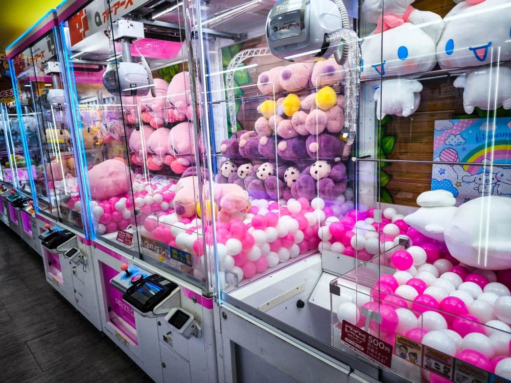 Eine Reihe von Spielautomaten, gefüllt mit Plüschtieren in Rosa, Lila und Weiß, umgeben von rosa und weißen Plastikbällen, in einer hell erleuchteten Spielhalle am Nakano Broadway.