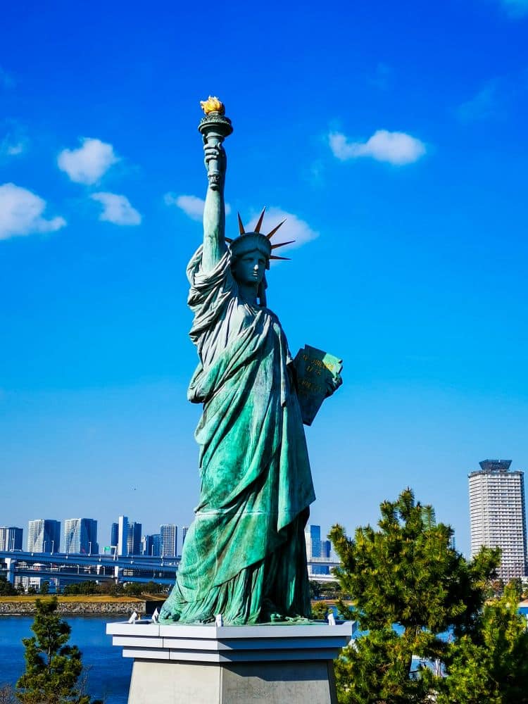 Die Freiheitsstatue erhebt sich vor einem strahlend blauen Himmel mit der Skyline von New York City und dem Wasser im Hintergrund, eingerahmt von grünen Bäumen im Vordergrund.