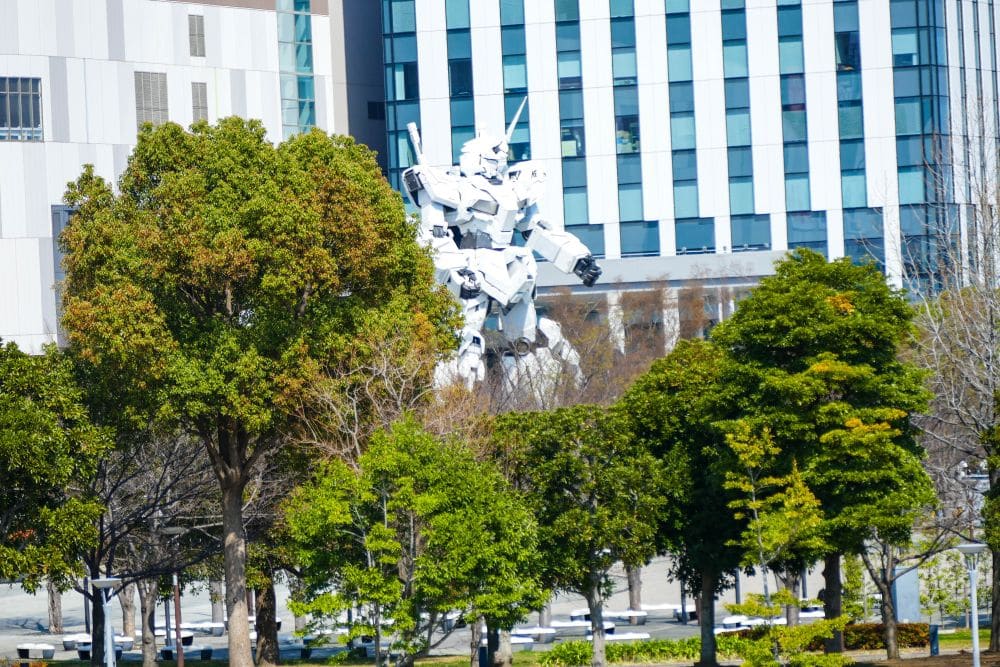 Eine große weiße Unicorn-Gundam-Roboterstatue ist teilweise hinter grünen Bäumen in Odaiba zu sehen, mit hohen modernen Gebäuden im Hintergrund unter einem klaren Tokioter Himmel.