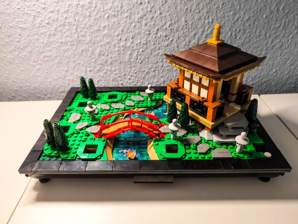 Ein LEGO Modell zeigt einen japanischen Garten mit einer roten Bogenbrücke über einen blauen Bach, üppiger grüner Bepflanzung, Steinlaternen und einem braunen und hellbraunen Gebäude im traditionellen Stil auf einem rechteckigen Sockel.