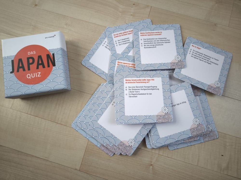Eine Schachtel mit der Aufschrift "Das Apan-Quiz" steht auf einem Holzboden neben mehreren ausgebreiteten Japan-Quiz-Karten mit gemusterten Rückseiten und deutschem Text, die jeweils eine Überschrift, Aufzählungspunkte und einen rot hervorgehobenen Titel enthalten.