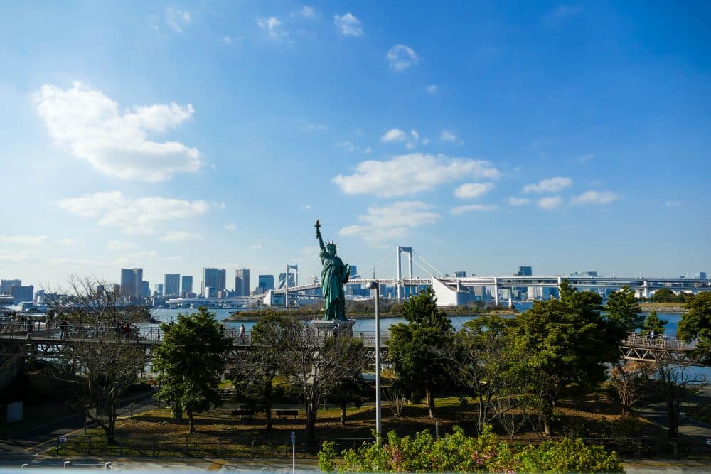 Eine Nachbildung der Freiheitsstatue von Tokio steht in einem Park mit Bäumen und Blick auf das Wasser und die Skyline der Stadt, die an New York erinnert, mit hohen Gebäuden und einer großen Brücke unter einem strahlend blauen Himmel mit vereinzelten Wolken.
