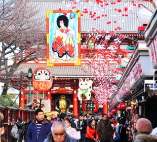Eine belebte Straße in Japan zu Hatsumōde, die mit Laternen, roten und weißen Ornamenten und großen Schildern mit traditionellen Kunstwerken, einer Daruma-Puppe, einer Glückskatze und Geschäften auf beiden Seiten geschmückt ist.
